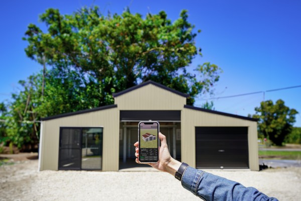 A person's arm holding a phone with the FDB Shed Designer App open positioned in front of a real-life shed matching the design on screen