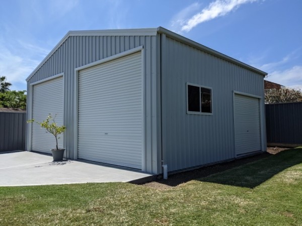 Double light grey garage with a gable roof and two white roller doors at the front, plus one extra roller door to the right side. A potted plant is on the driveway to the left between the roller doors.
