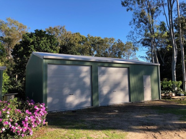 A green garage with white doors in a garden setting with purple flowers in the front left